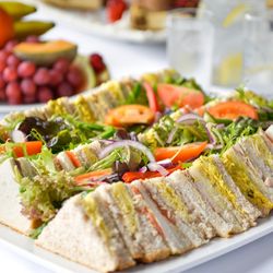 A platter of assorted tea sandwiches with various fillings, garnished with fresh lettuce, tomato slices, and red onion, served on a white plate with fruit and water in the background.