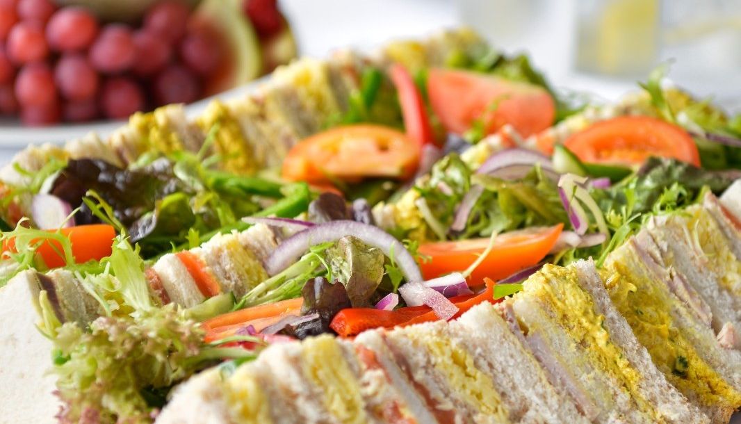 A platter of assorted tea sandwiches with various fillings, garnished with fresh lettuce, tomato slices, and red onion, served on a white plate with fruit and water in the background.