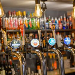 Close-up of beer taps at a bar, featuring brands like Strongbow, Amstel, John Smith's, and Foster's, with shelves of liquor bottles and hanging light bulbs in the background.