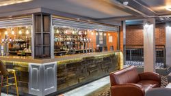 Modern bar interior with a wooden counter, leather chairs, exposed brick walls, and shelves stocked with various bottles and taps for drinks.