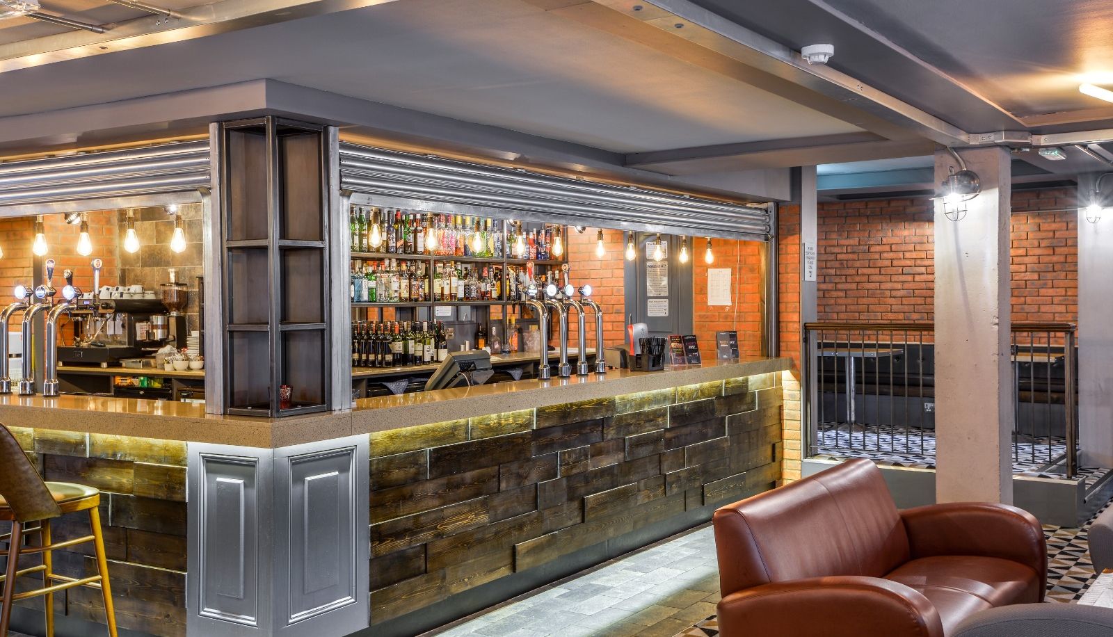 Modern bar interior with a wooden counter, leather chairs, exposed brick walls, and shelves stocked with various bottles and taps for drinks.