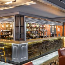 Modern bar interior with a wooden counter, leather chairs, exposed brick walls, and shelves stocked with various bottles and taps for drinks.