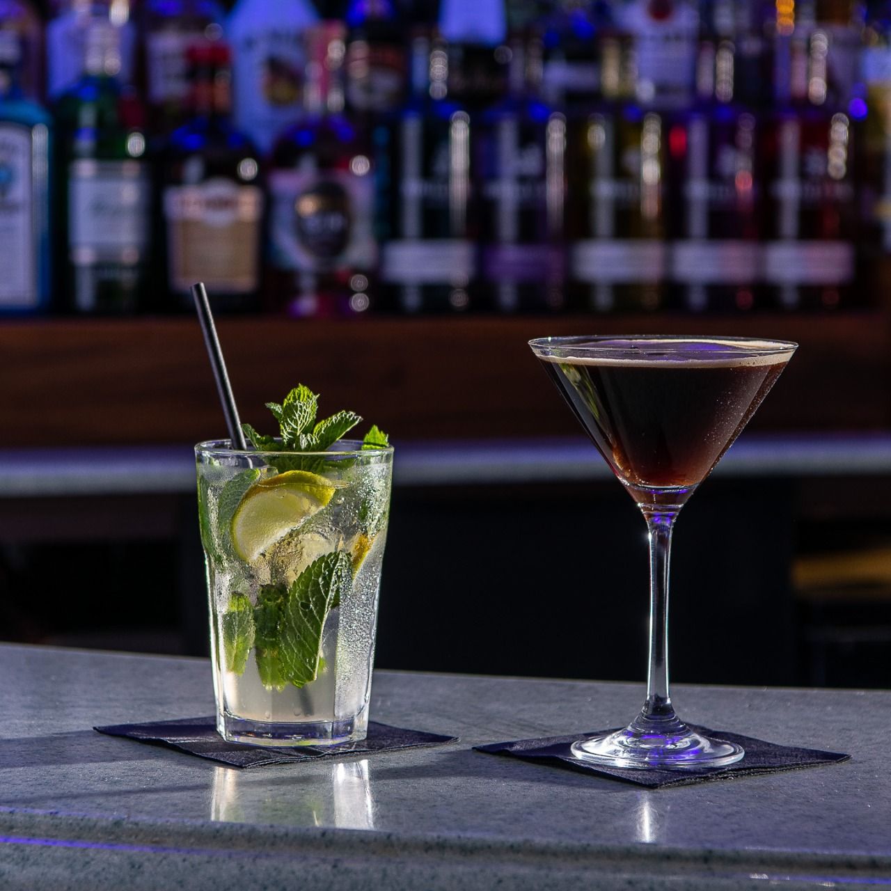 Two cocktails on a bar counter, one with mint and lime in a highball glass and the other in a martini glass, with bottles in the background.