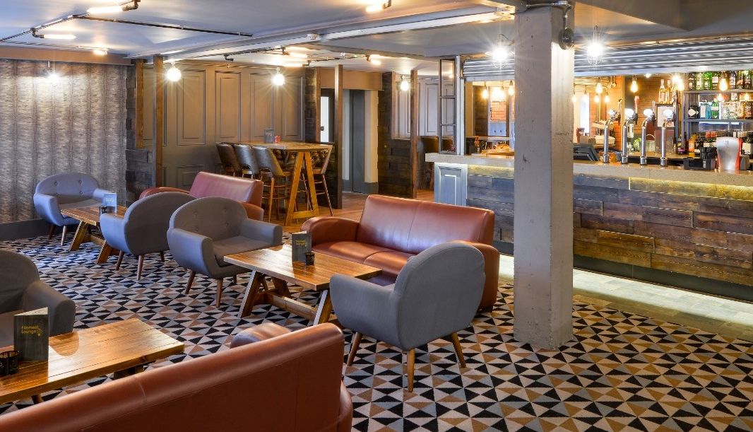 Interior of a modern bar with geometric patterned carpet, brown leather sofas, grey armchairs, wooden tables, and a well-stocked bar counter in the background.