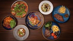A top view of a variety of dishes on a wooden table, including a pizza with arugula, pasta dishes, steaks served with fries and vegetables, and a plate with grilled chicken and sauce.
