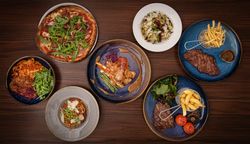A top view of a variety of dishes on a wooden table, including a pizza with arugula, pasta dishes, steaks served with fries and vegetables, and a plate with grilled chicken and sauce.