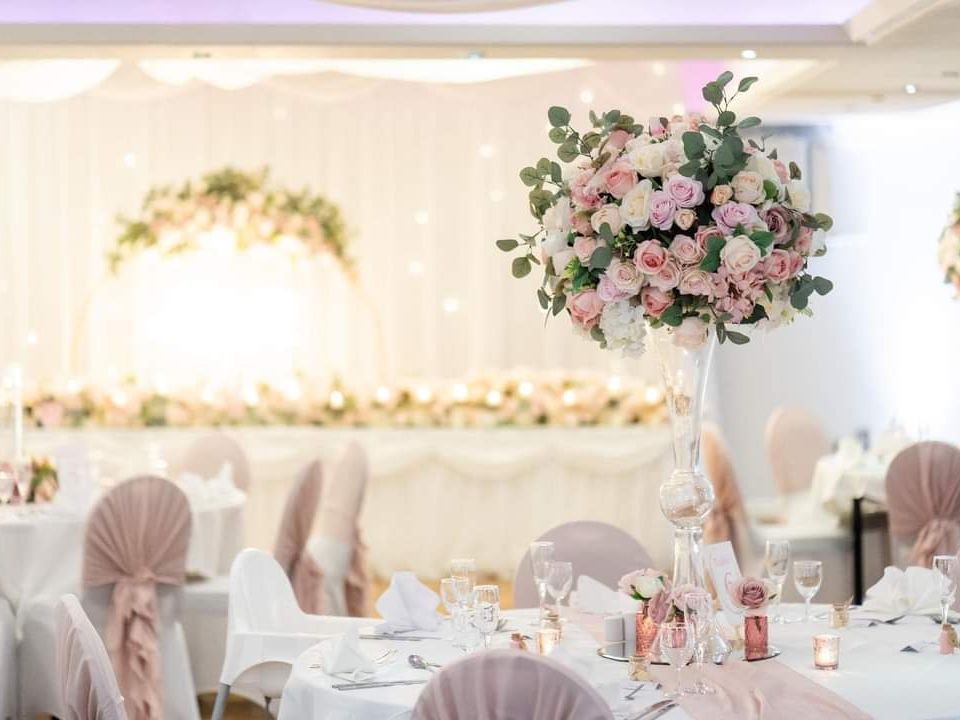 Elegant wedding reception with round tables decorated with pink floral centerpieces, soft pink chair covers, and delicate candles in a bright, romantic setting.