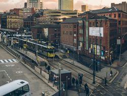 Urban cityscape at sunset featuring a tram, double-decker bus, pedestrians, and a mix of modern and historic buildings.