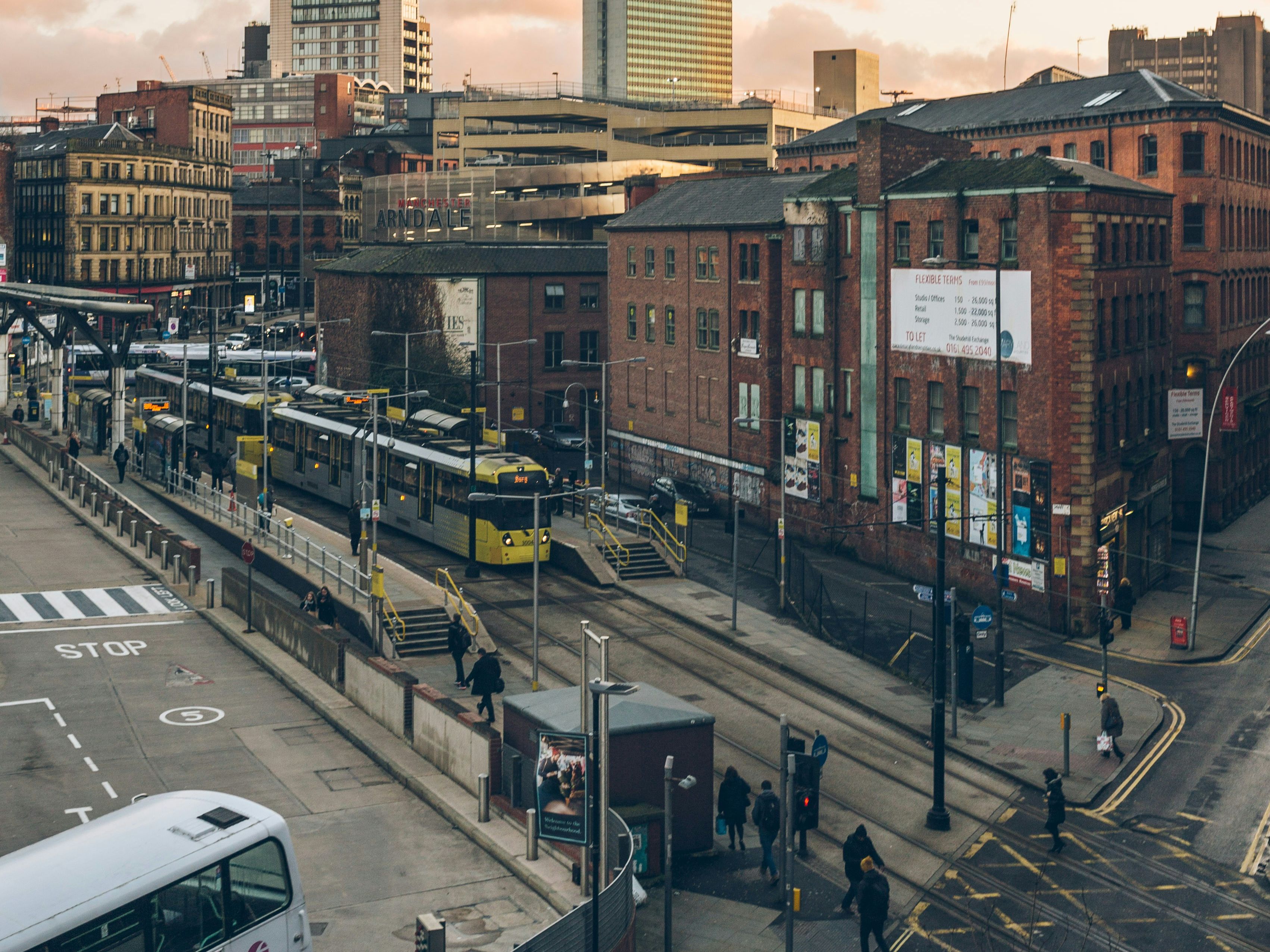 Urban cityscape at sunset featuring a tram, double-decker bus, pedestrians, and a mix of modern and historic buildings.