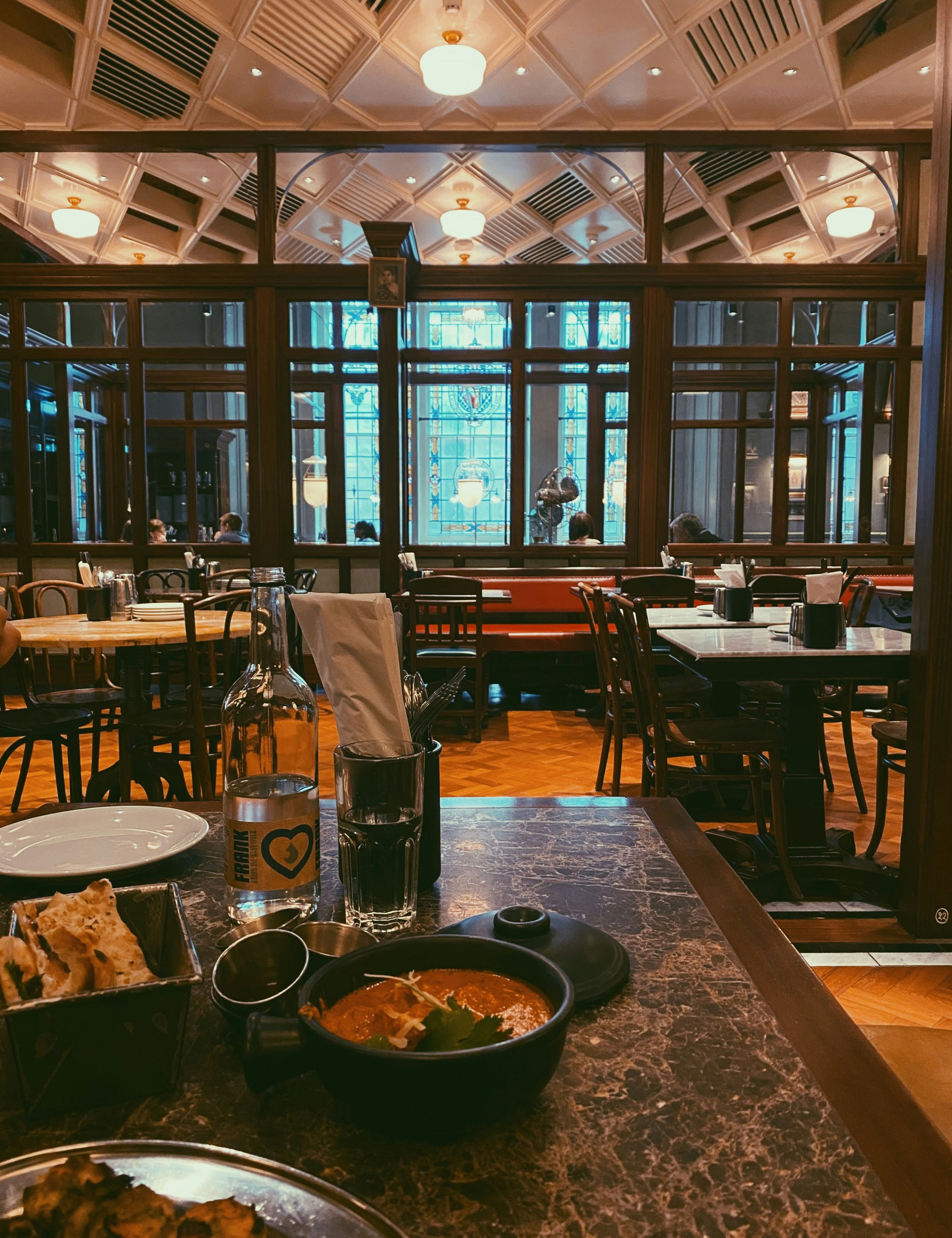 A cozy restaurant with marble tables, wooden chairs, and large stained glass windows. There is a bowl of curry, naan bread, a glass of water, and a bottle on the table in the foreground.