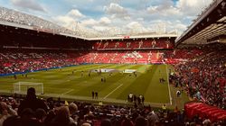 View of a crowded football stadium with fans in the stands and players warming up on the pitch, under a partly cloudy sky.