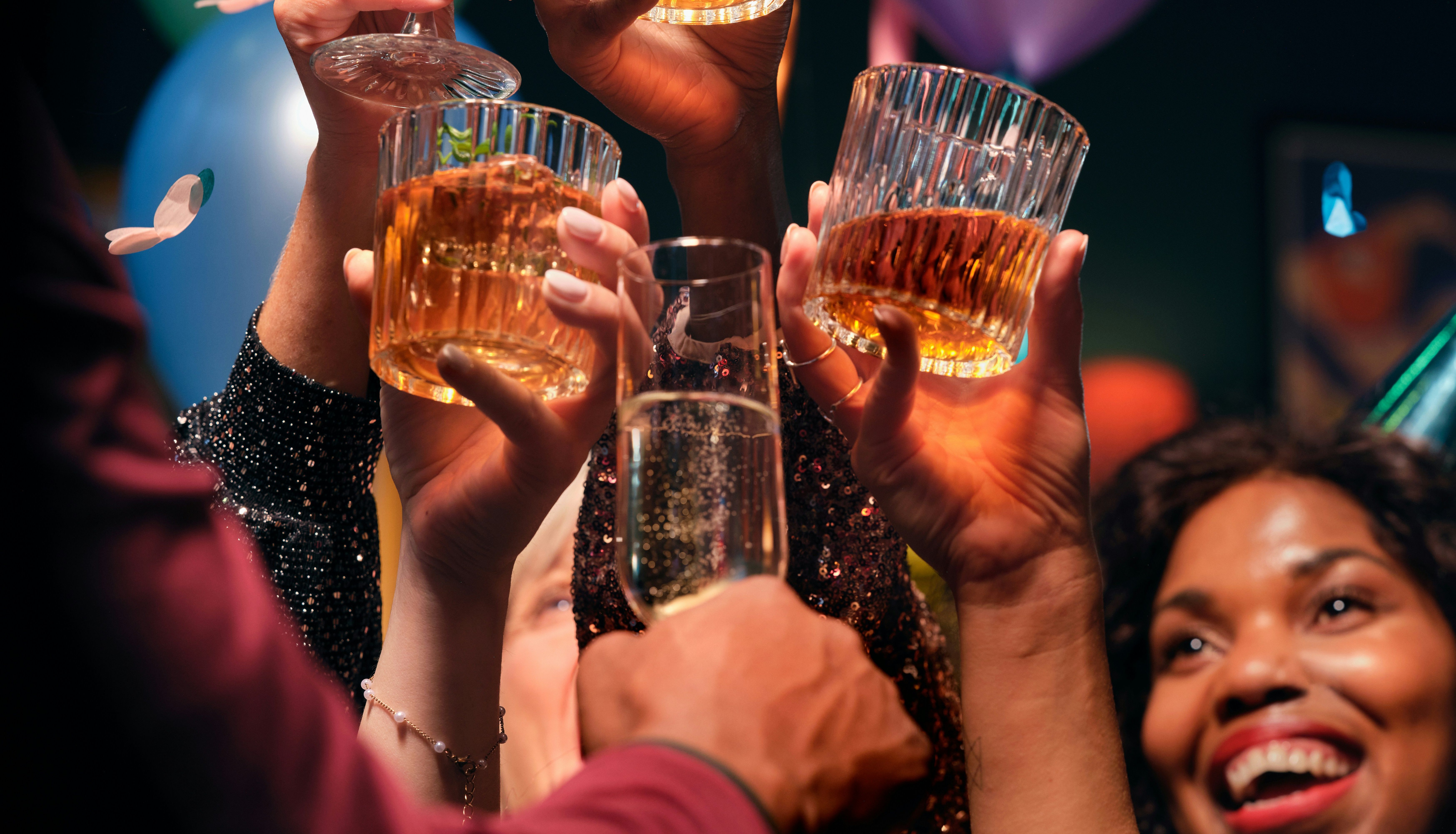 Group of people celebrating and toasting with drinks at a festive party with balloons and confetti.