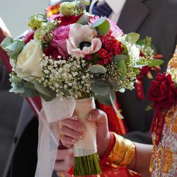 Close-up of a hand holding a colorful bridal bouquet with white, pink, and red flowers, in a wedding setting with traditional Chinese attire and gold jewelry.