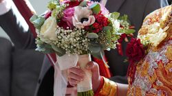 Close-up of a hand holding a colorful bridal bouquet with white, pink, and red flowers, in a wedding setting with traditional Chinese attire and gold jewelry.