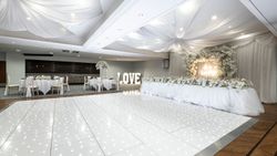 Elegant wedding reception hall decorated in white with flowers, draped fabric ceiling, a long head table, illuminated dance floor, and large 'LOVE' letters.
