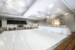 Elegant wedding reception hall decorated in white with flowers, draped fabric ceiling, a long head table, illuminated dance floor, and large 'LOVE' letters.
