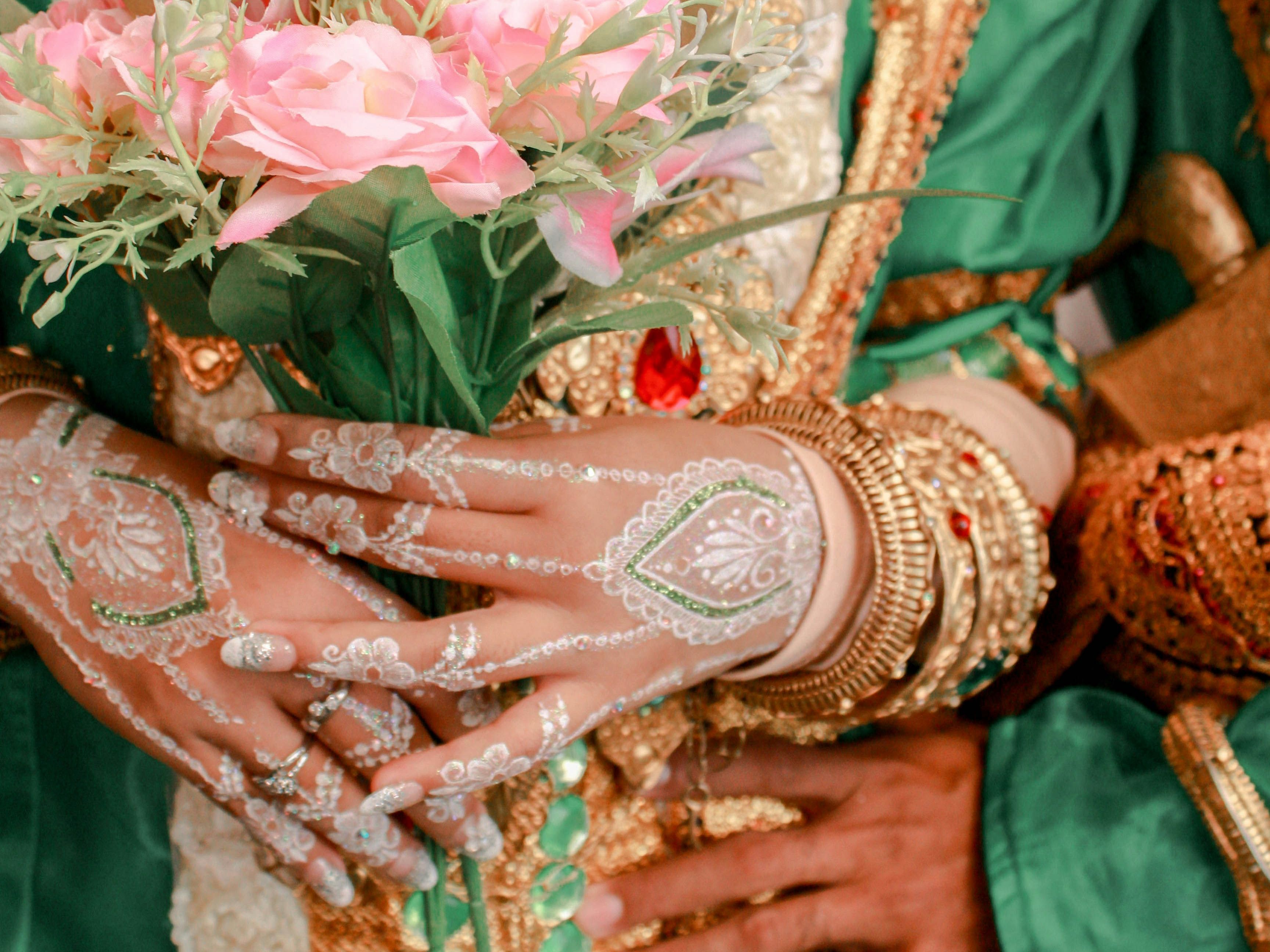 Close-up of hands with intricate white and green henna holding a bouquet of pink flowers, adorned with gold jewelry and wearing traditional green and gold attire.