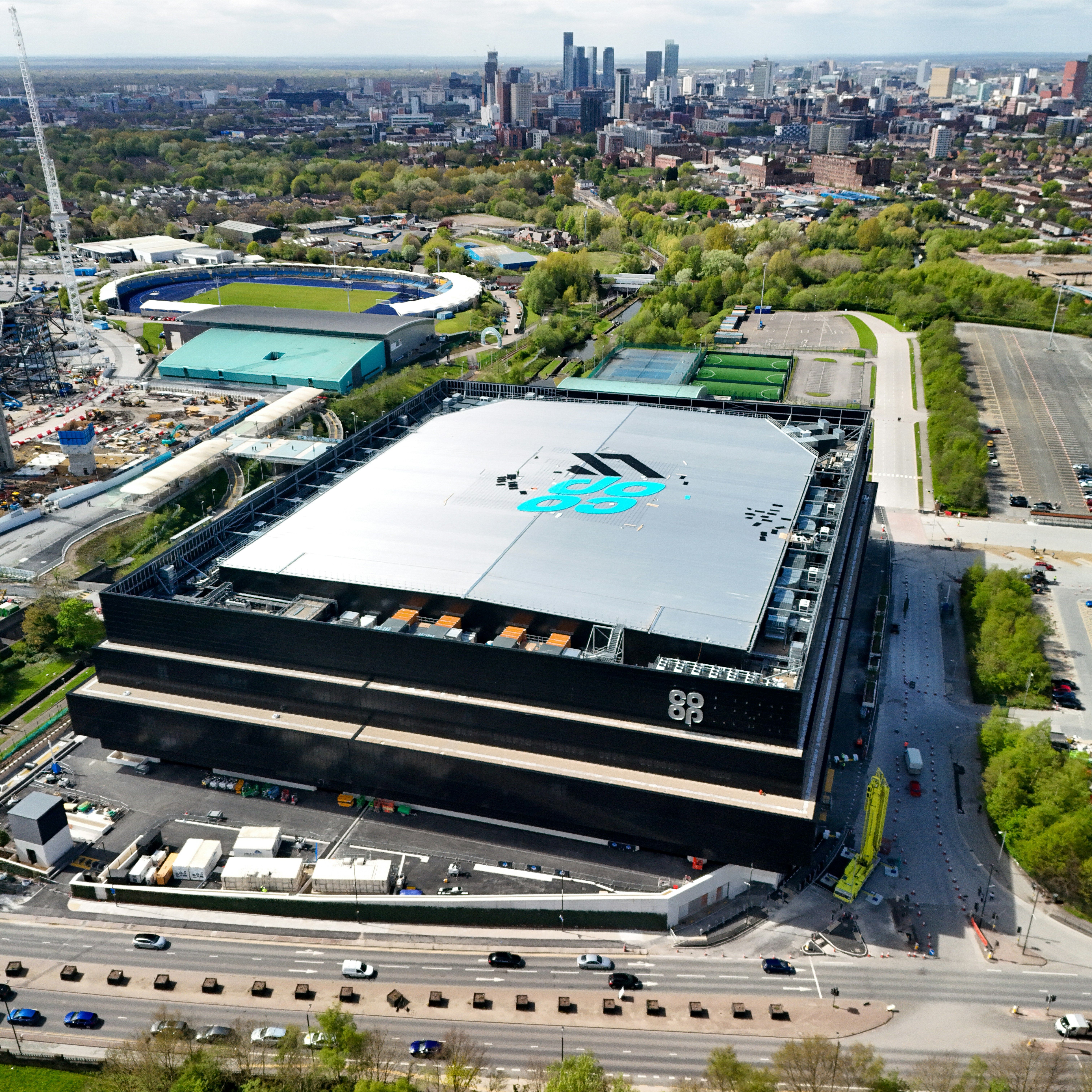 Aerial view of the Co-op Live arena under construction in Manchester, with the city skyline in the background and nearby sports facilities visible.