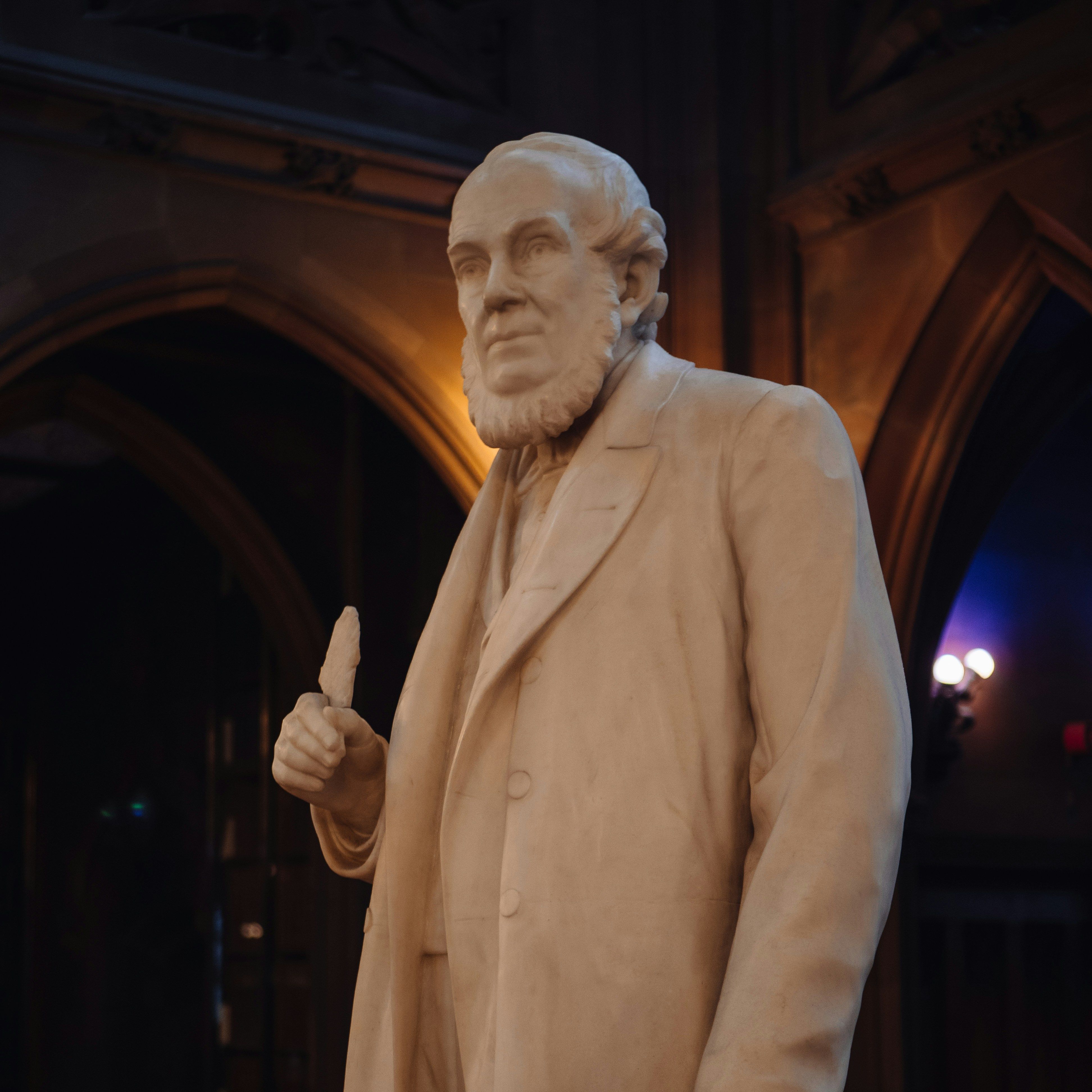 Marble statue of a bearded man in a suit holding an object, standing indoors with ornate architectural details in the background.