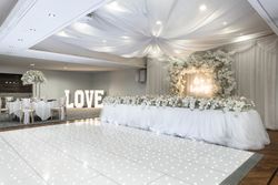 Elegant wedding reception venue decorated in white, featuring draped ceilings, a sparkling dance floor, floral arrangements, a long flower-adorned table, and illuminated 'LOVE' letters.