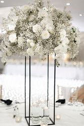 Elegant floral centerpiece with white roses, baby's breath, and white orchids arranged on a tall black metal stand, with floating candles in glass vases on a white table.