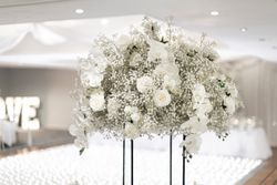 Elegant floral arrangement centerpiece with white roses, orchids, and baby's breath at a wedding reception with a blurred 'LOVE' sign in the background.