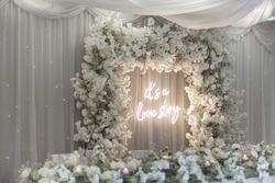 A romantic wedding backdrop with white flowers and a neon sign that reads 'It's a love story', surrounded by draped white fabric and floral arrangements.
