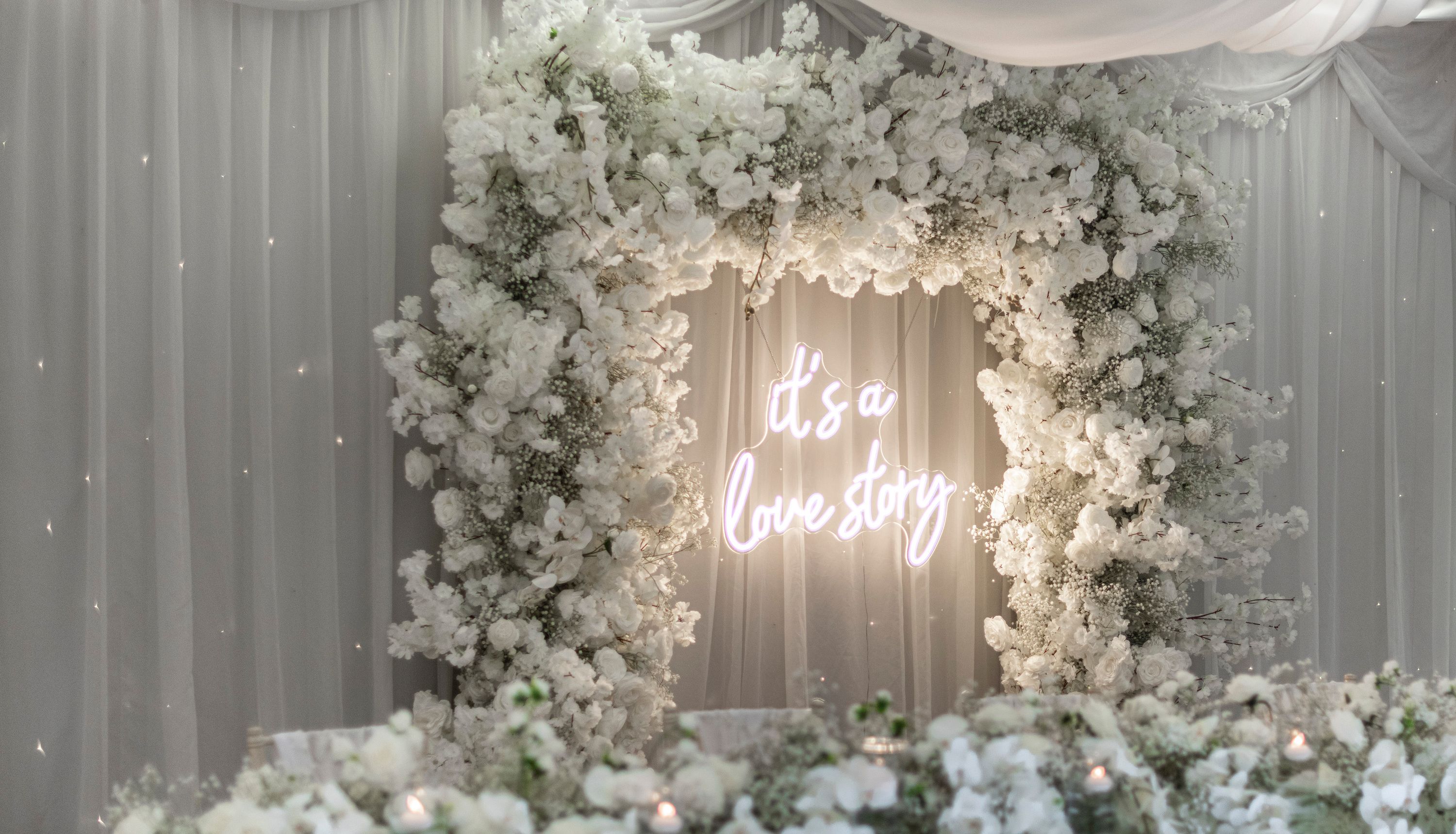A romantic wedding backdrop with white flowers and a neon sign that reads 'It's a love story', surrounded by draped white fabric and floral arrangements.