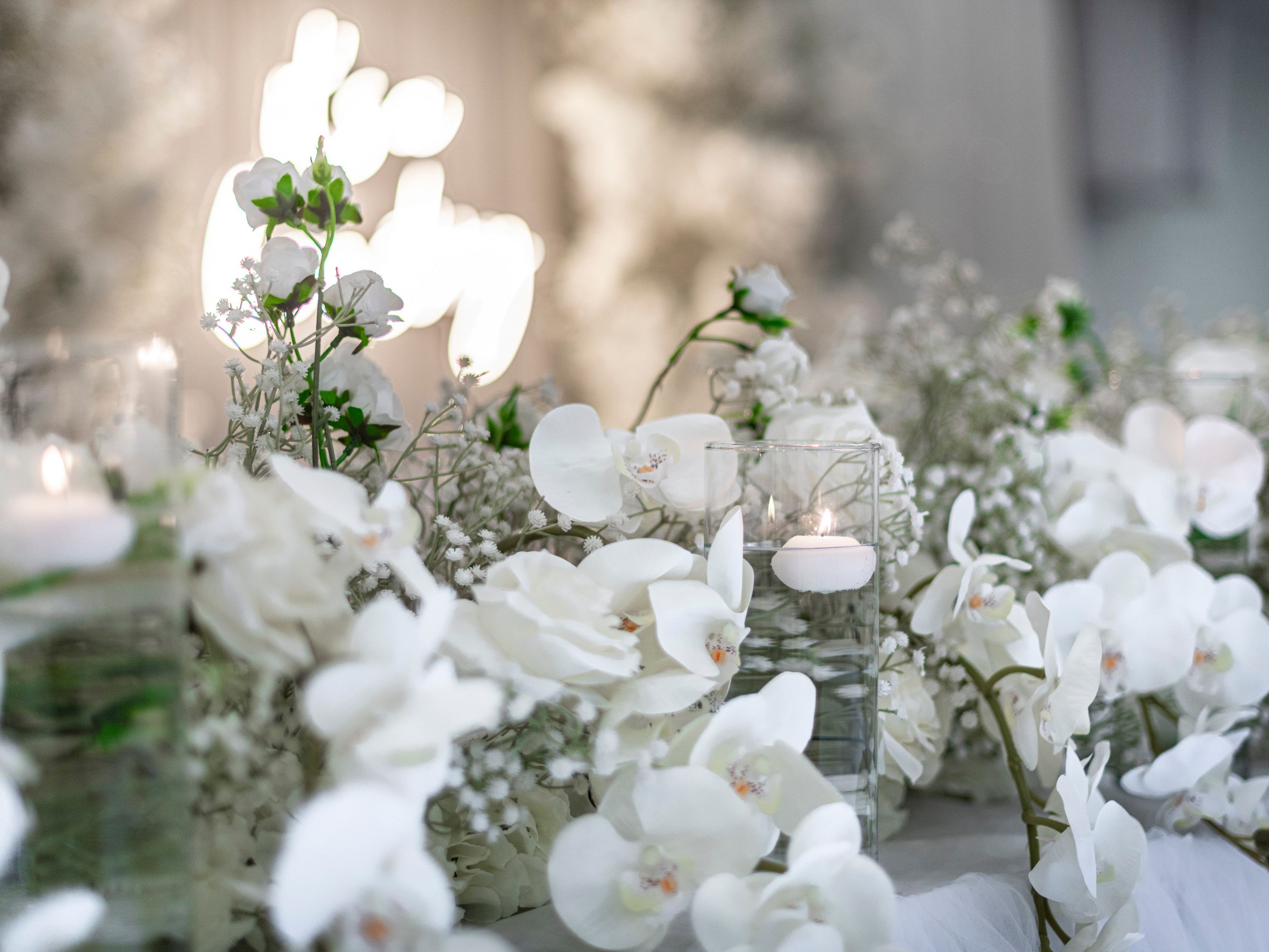 Elegant white floral arrangement with candles in glass holders, featuring orchids and baby's breath, set on a decorated table.