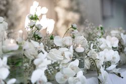 Elegant white floral arrangement with candles in glass holders, featuring orchids and baby's breath, set on a decorated table.