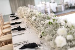 Elegant banquet table set for an event with white floral centerpieces, glassware, silver cutlery, white plates, and black napkins on a white tablecloth.
