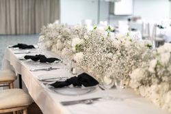Elegant wedding table setting with white floral centerpiece, glass plates, silver cutlery, black napkins, and soft beige chairs.