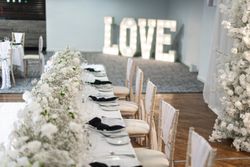 Elegant wedding reception table set with white flowers and black napkins, with illuminated 'LOVE' sign in the background.