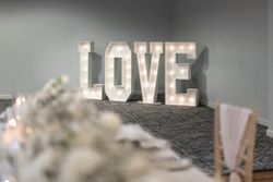 Large illuminated letters spelling 'LOVE' set up in a decorated indoor event space with a blurred foreground table setup.