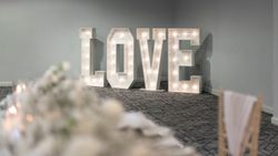 Large illuminated letters spelling 'LOVE' set up in a decorated indoor event space with a blurred foreground table setup.