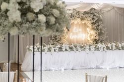 Elegant wedding reception setup with a white floral arrangement, draped fabric, and a neon sign that reads 'It's a love story'.