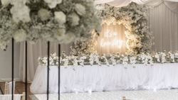 Elegant wedding reception setup with a white floral arrangement, draped fabric, and a neon sign that reads 'It's a love story'.