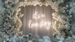 Neon sign that says 'it's a love story' surrounded by a large white floral arrangement, likely used as a wedding or romantic event backdrop.
