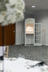 A white vintage telephone booth with a sign inside that reads 'Audio Guestbook,' set up in a modern indoor event space with a table and floral arrangement visible in the foreground.