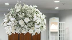 Large arrangement of white roses, baby's breath, and white orchids displayed indoors with a blurred background.
