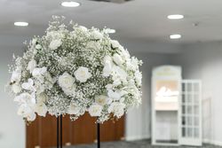 Large arrangement of white roses, baby's breath, and white orchids displayed indoors with a blurred background.