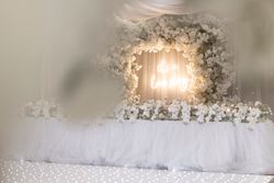 Elegant wedding decoration with a large arch of white flowers and a neon sign that reads 'it's a love story', complemented by white floral arrangements on a draped table.