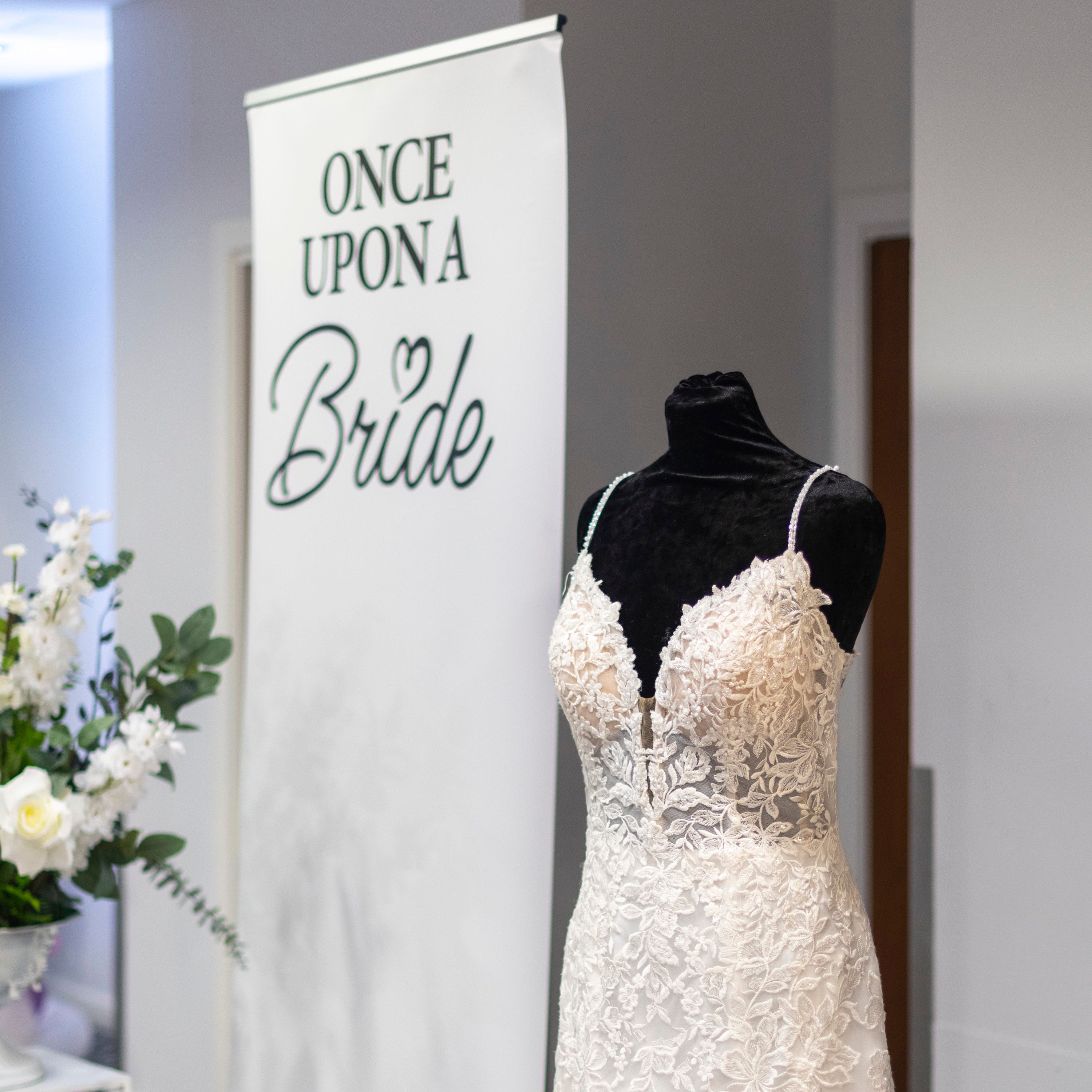 A white lace wedding dress on a black mannequin next to a sign that reads 'Once Upon A Bride' with flower arrangements in the background.