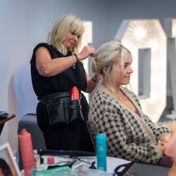 A hairstylist is doing a woman's hair in a salon setting, with large illuminated letters spelling 'LOVE' in the background, and hair styling products on a table in the foreground.