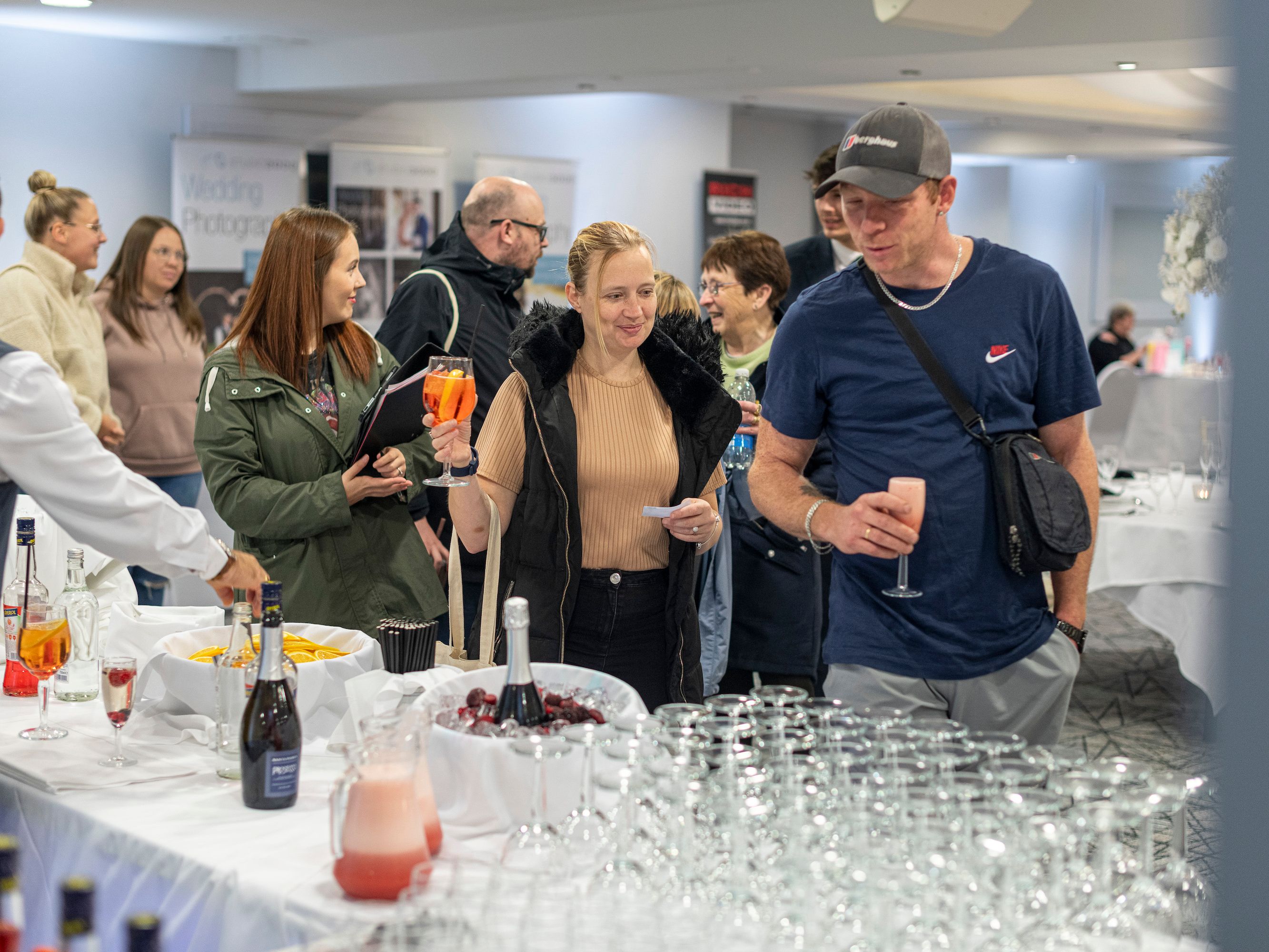 People gathered around a table with various drinks at a social event, some holding glasses and interacting with each other.
