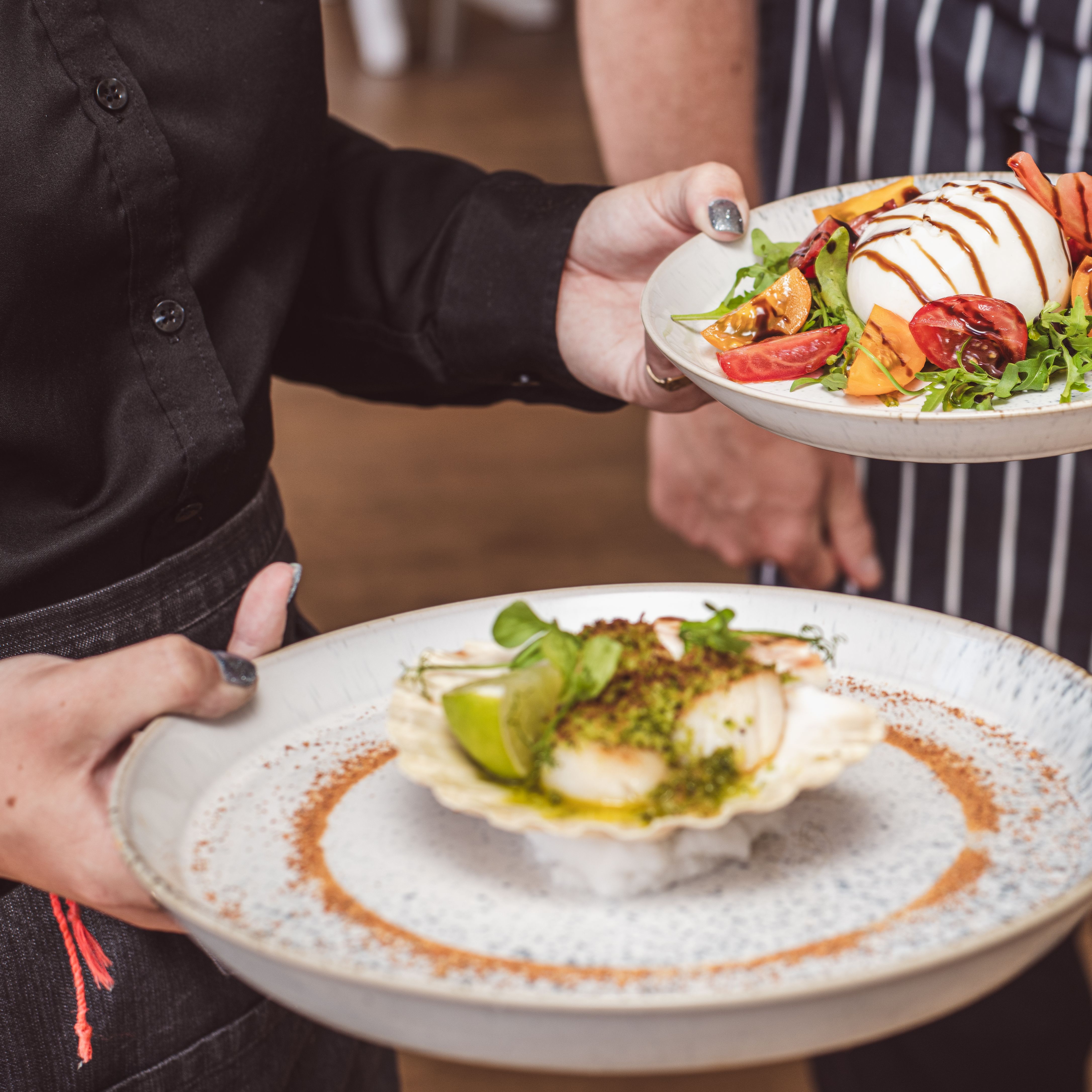 Waiter carrying plated dishes including burrata with tomatoes and a scallop starter