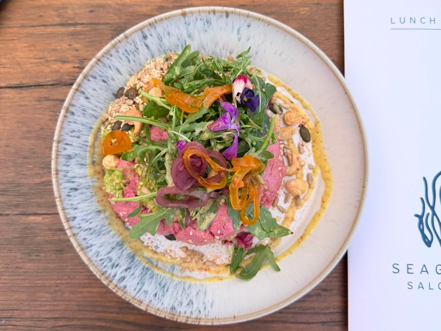 Colourful salad bowl with rocket, pickled onions and edible flowers on a wooden table beside a lunch menu