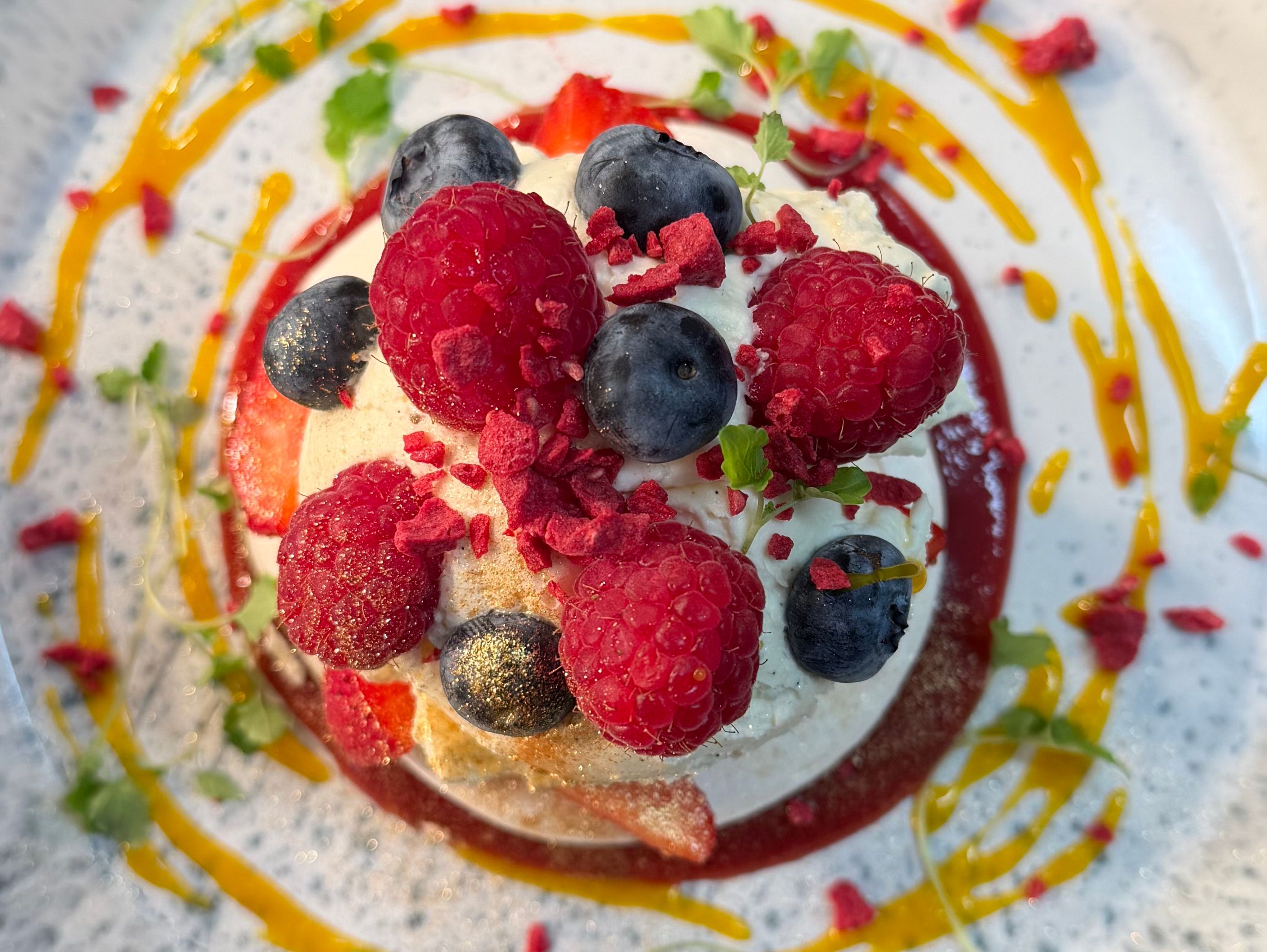 Berry dessert topped with raspberries and blueberries, drizzled with fruit sauce on a white speckled plate