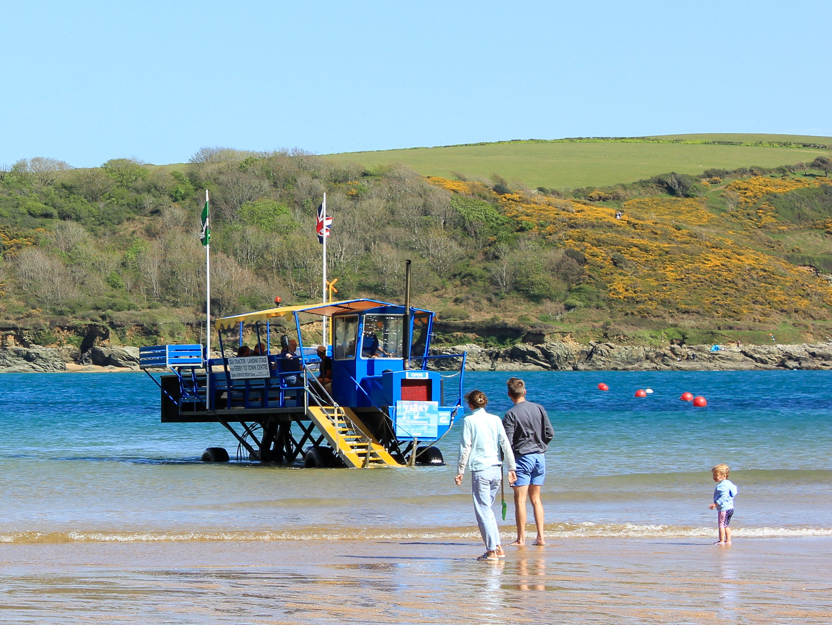 People on a sandy beach near a unique blue passenger vehicle on stilts in shallow water, with grassy hills in the background.
