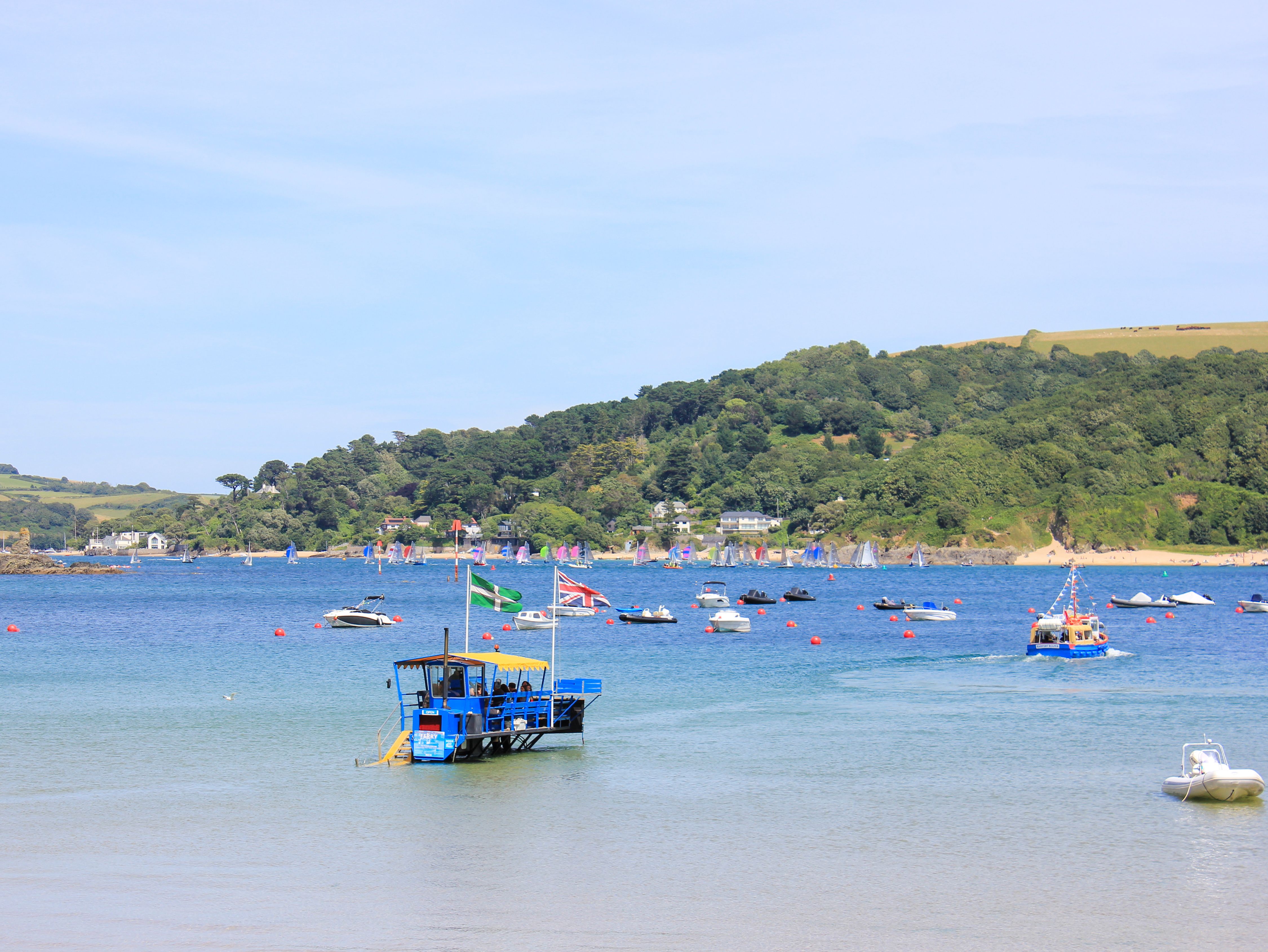 Boats floating on a calm blue estuary with green hills and scattered houses in the background.